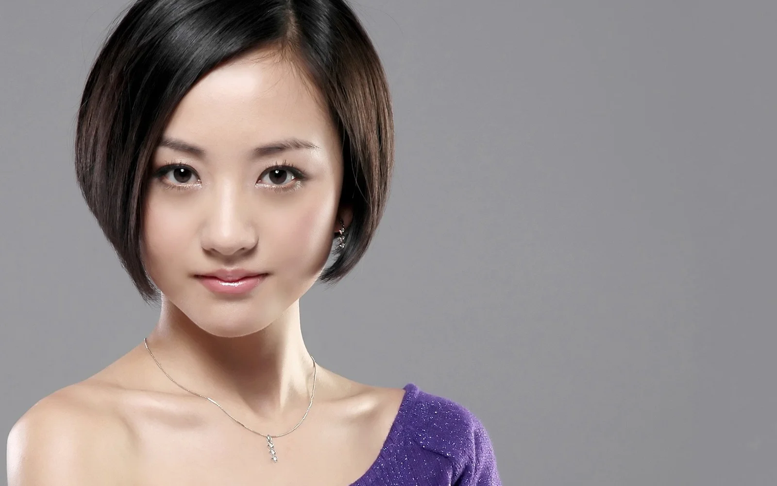 一名短发女性面对镜头，身穿紫色上衣，背景为纯灰色。