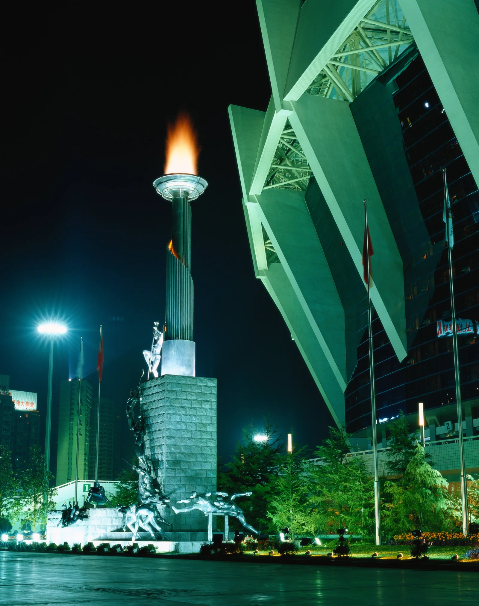 夜色中的城市广场矗立着灯火通明的纪念碑，旁边是一座现代建筑。