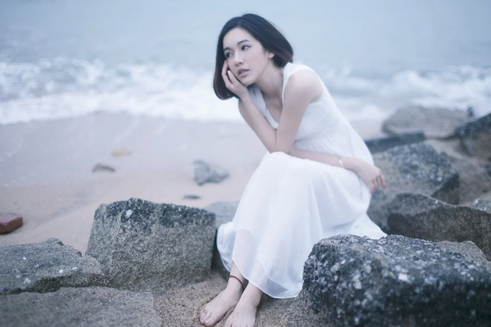一名身穿白裙的成年女性坐在海邊岩石上。