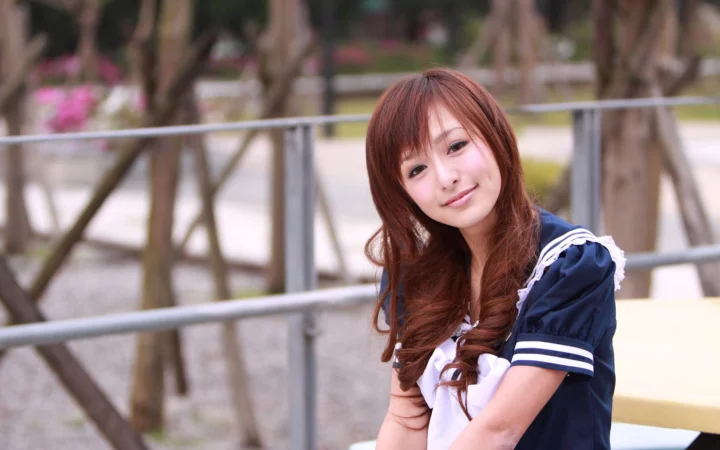 茶色の長い髪の女性が紺色のセーラー風トップスを着て、木々と手すりのそばでカメラに微笑みかけています。