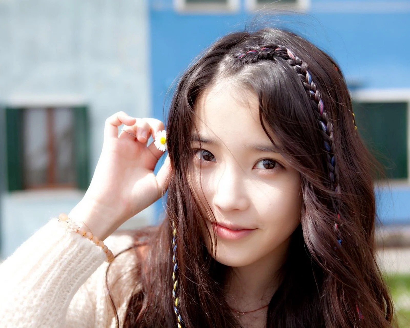 땋은 머리의 여성이 야외에서 작은 꽃을 들고 햇살 아래 카메라를 향해 미소 짓고 있다.