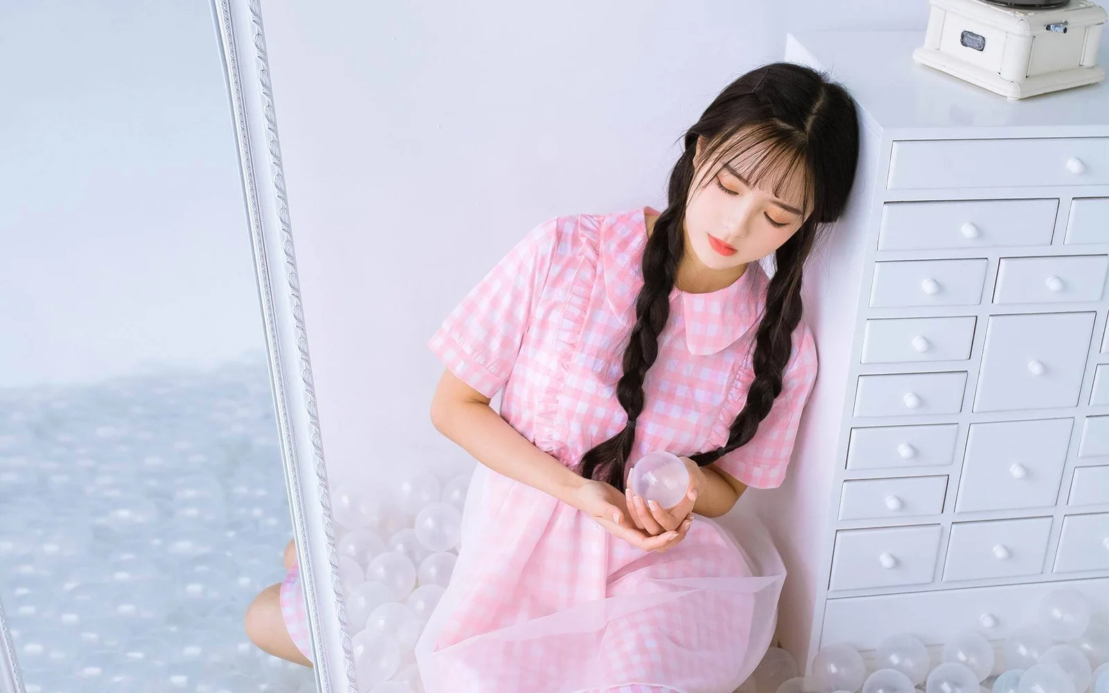 一名梳着双辫、身穿粉色格纹连衣裙的女性靠在白色抽屉柜旁，手里拿着一个小球。
