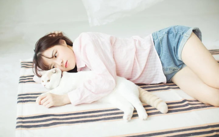 一名穿粉色上衣和牛仔短裤的女子躺在条纹床单上，身旁是一只白猫。