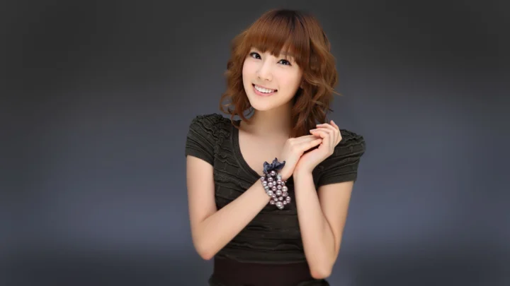 검은색 상의를 입은 성인 여성이 짙은 회색 배경 앞에서 미소를 지으며 포즈를 취하고 있습니다.