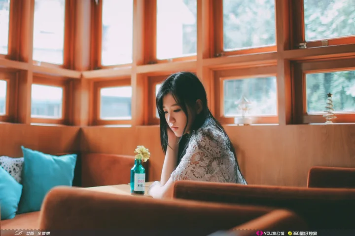 一名女子坐在明亮的窗边室内空间中，身旁是沙发和一瓶小花。
