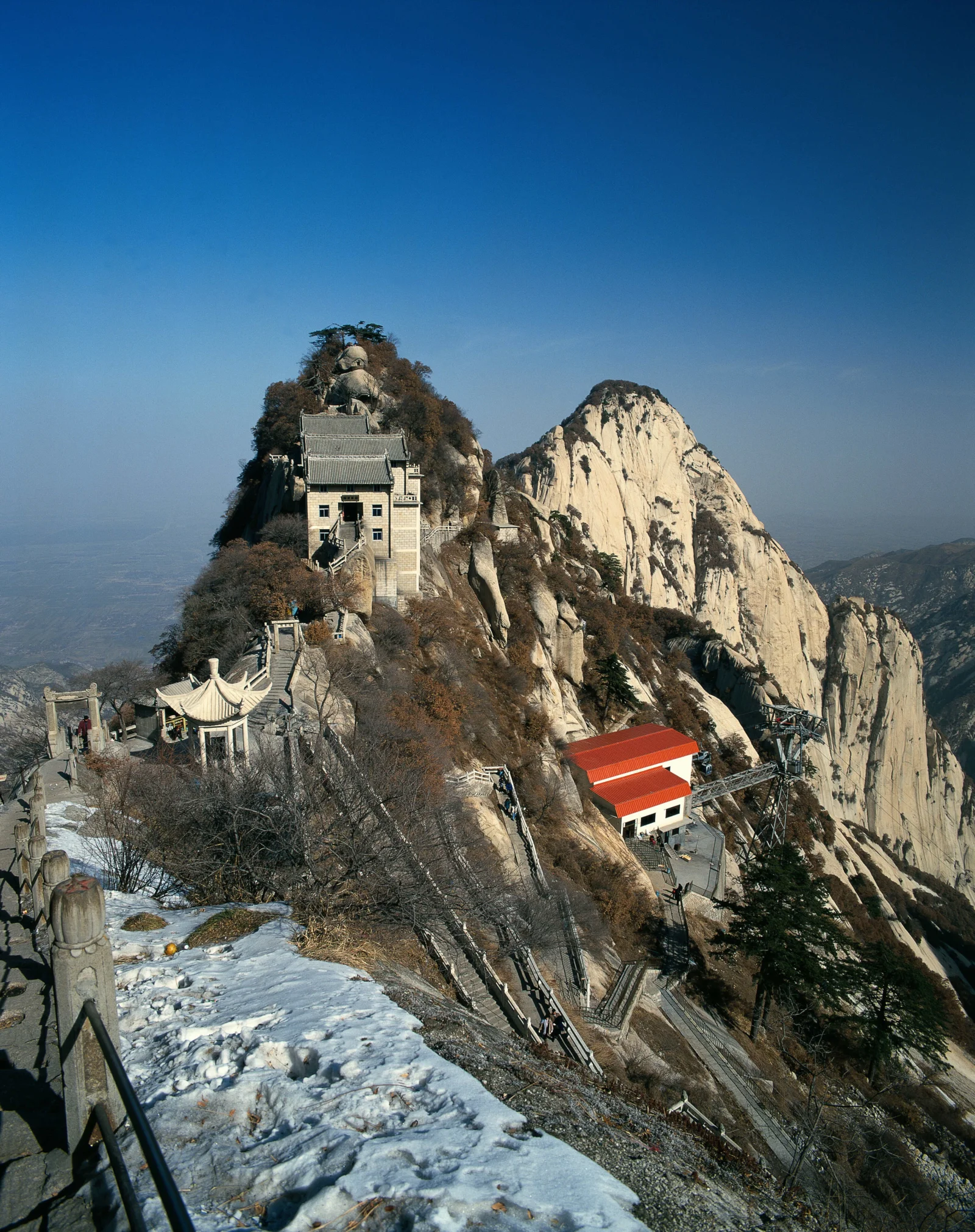 急峻な岩壁と雪道の間に寺院が建ち、遠くに連なる山々が広がっている。