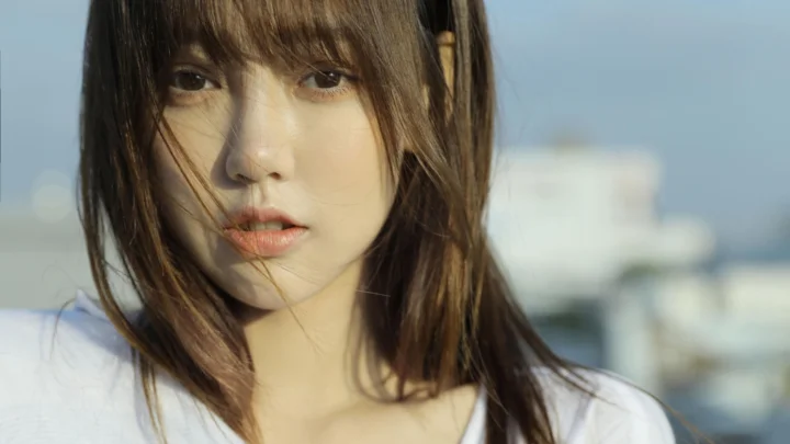바람에 머리카락이 흩날리는 갈색 머리 여성이 흐릿한 도시 풍경을 배경으로 카메라를 바라보고 있다.