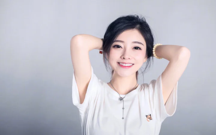 一名身穿白色上衣的微笑女性在浅灰色背景前抬起双臂摆姿势。