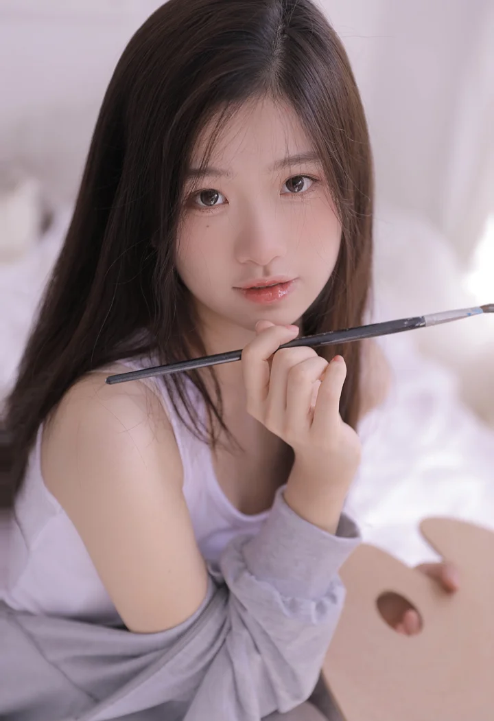 一名长发女性手持画笔，坐在柔和明亮的室内环境中。