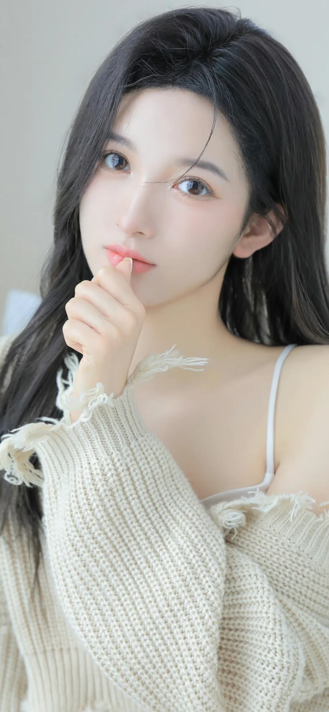 一名长发女性穿着米白色针织衫，手指轻触嘴唇面对镜头。
