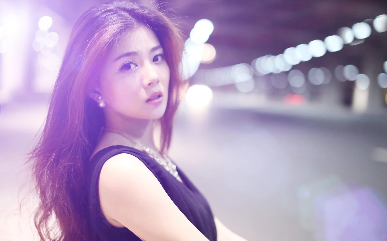 검은색 민소매 상의를 입은 여성이 밤의 흐릿한 도시 불빛을 배경으로 카메라를 바라보고 있다.