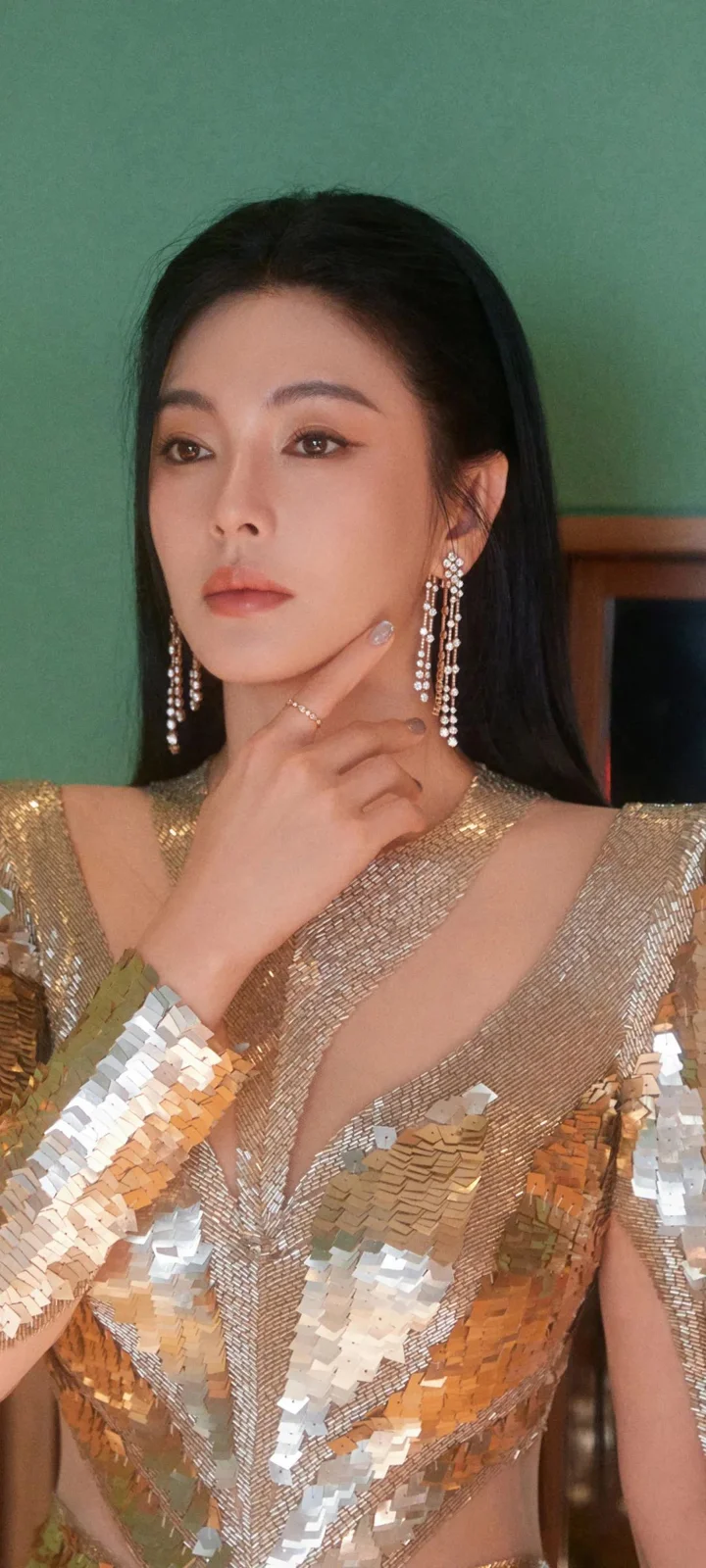 一位成年女性身穿金色亮片礼服，手指轻触下巴，在绿色墙面前面向镜头。