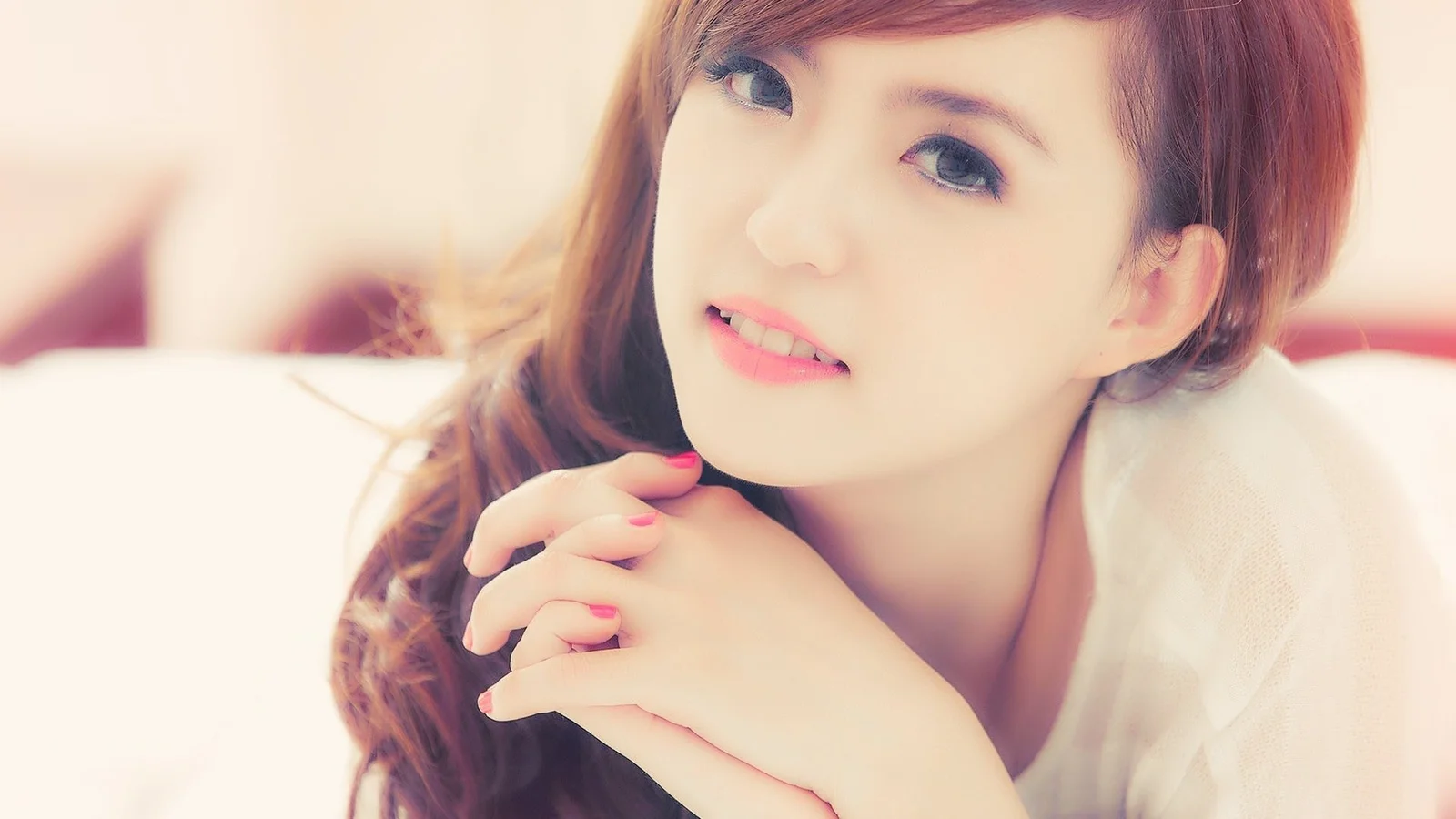 一位棕色长发女子在柔和光线下微笑着将双手叠放在脸旁。