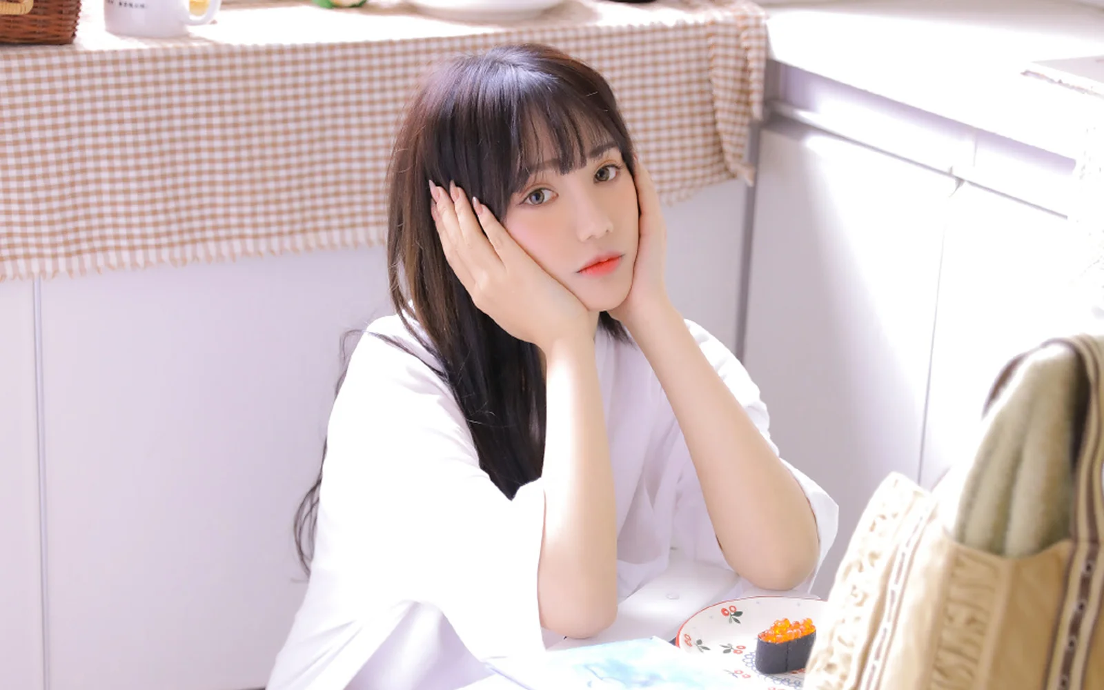 長い髪の女性が室内のテーブルのそばに座り、両手で頬を支えながらカメラを見ている。