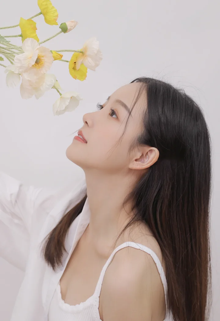 흰색 상의를 입은 긴 머리 여성이 옆모습으로 올려다보고 있으며 왼쪽 위에 흰색과 노란색 꽃이 보입니다.