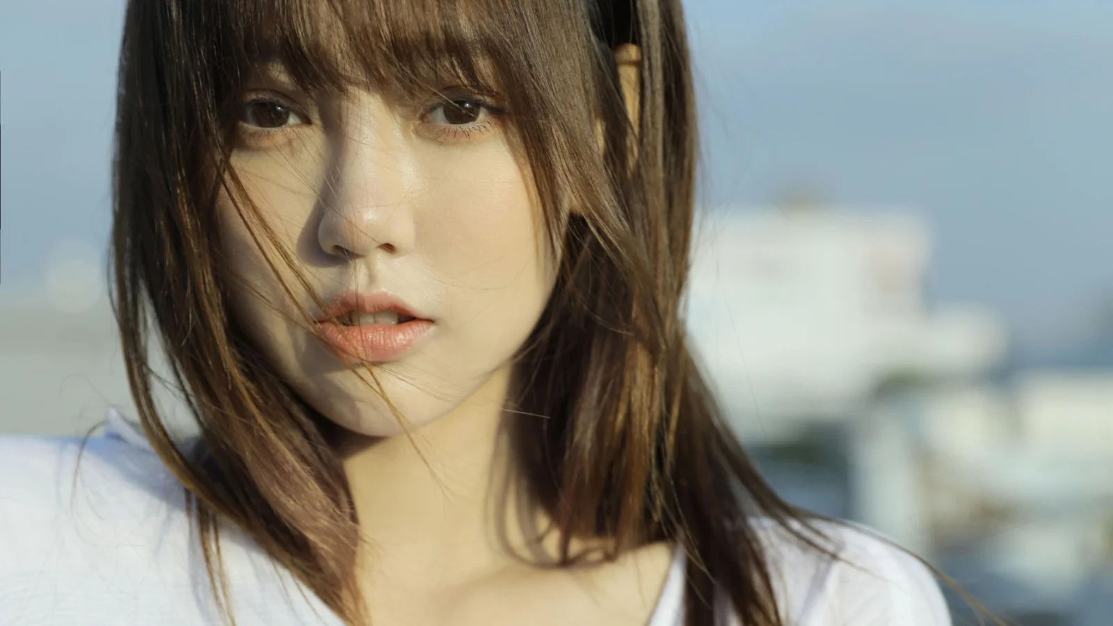 바람에 머리카락이 흩날리는 갈색 머리 여성이 흐릿한 도시 풍경을 배경으로 카메라를 바라보고 있다.