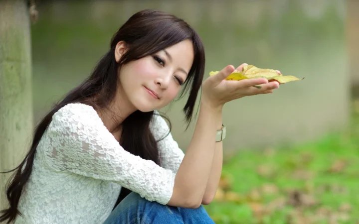 一名长发女子穿着白色上衣坐在户外，手中托着几片叶子。