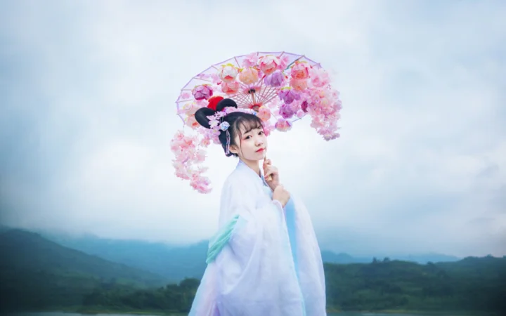 一名女子手持粉色花伞站在朦胧山景前。