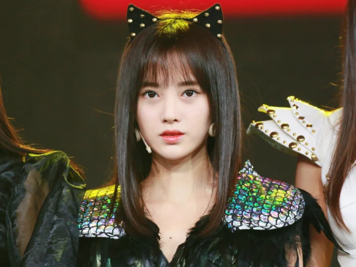 고양이 귀 머리띠와 반짝이는 깃털 장식을 한 의상의 여성을 정면에서 담은 인물 사진입니다.