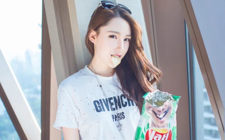 一名长发女性站在窗边，嘴里叼着薯片，手里拿着一袋薯片。