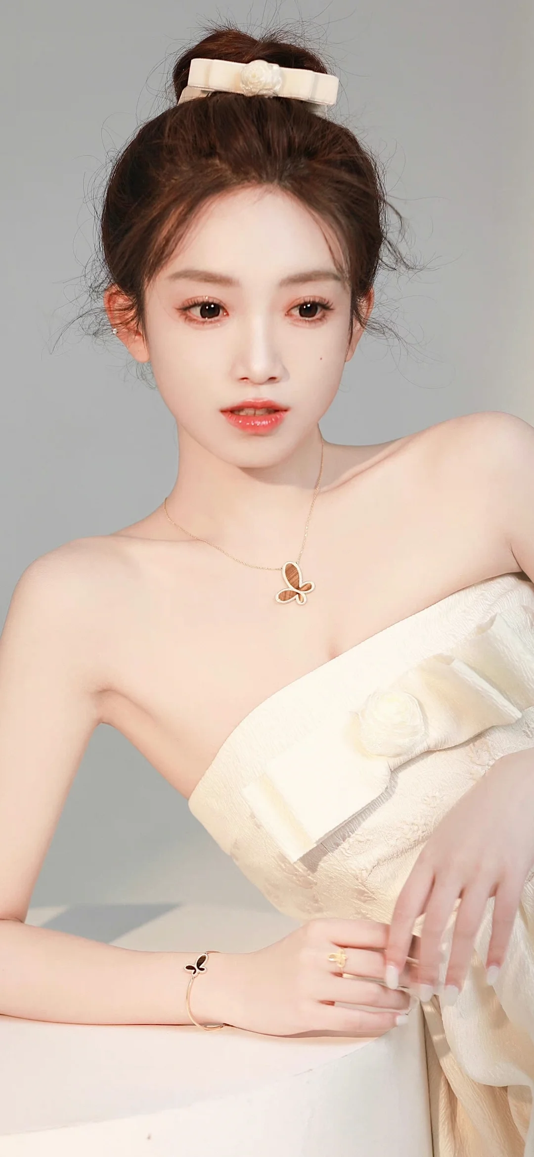 一位盘发女性身穿浅色无肩带礼服，面对镜头坐在浅灰色背景前。