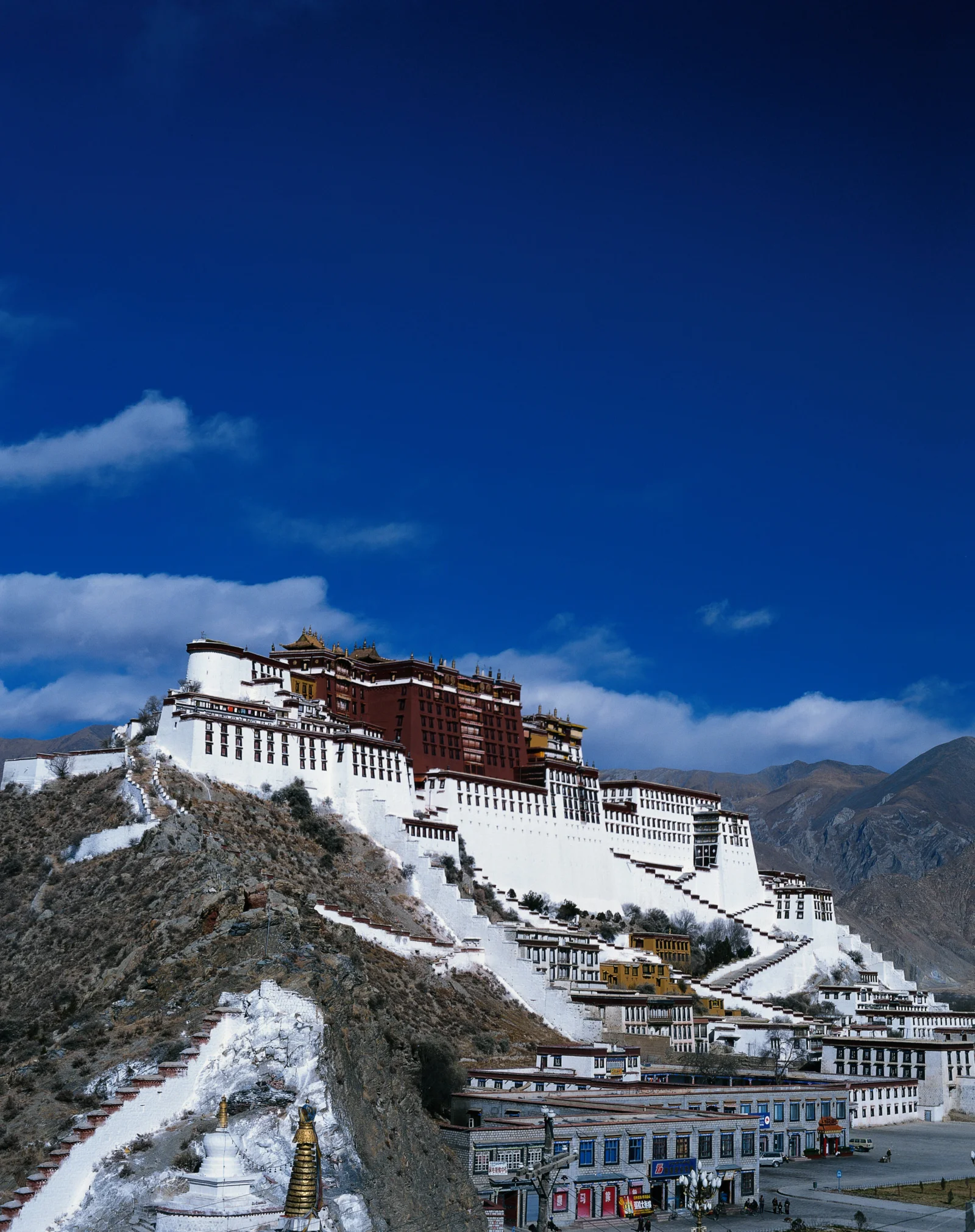 蓝天下的白色宫殿建筑坐落在覆雪山坡上，远处是群山。