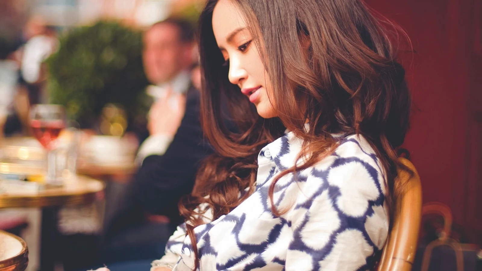 一名棕色长发女性身穿图案上衣坐在室内，侧头低头看向一侧。