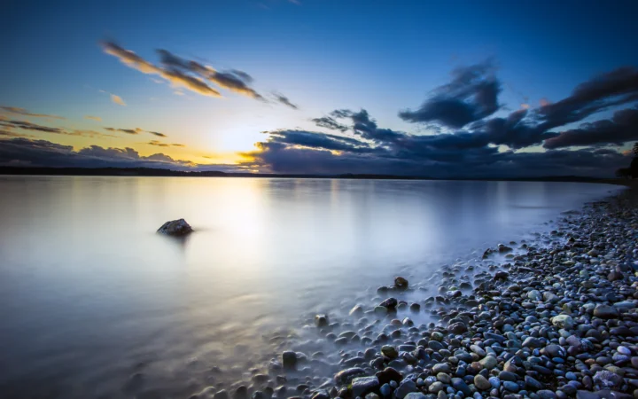 夕阳在平静湖面上投下金色倒影，远处乌云铺展，右侧是卵石岸边。