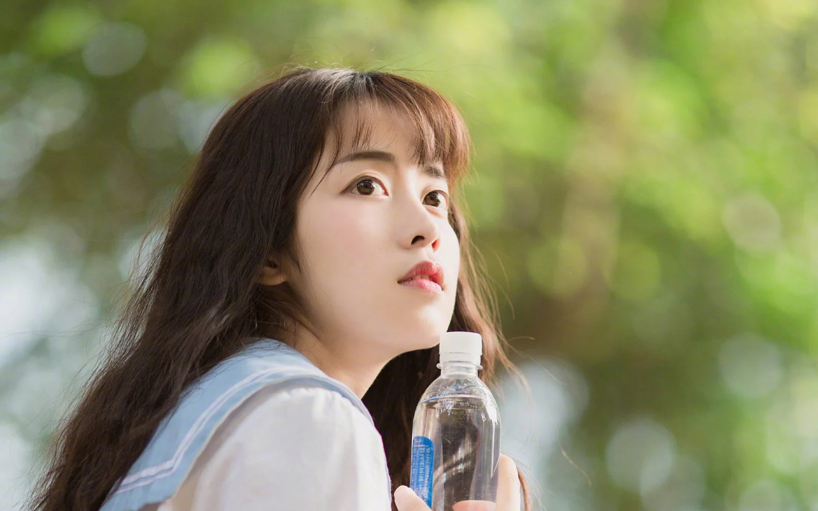 一名女子在绿色户外背景前侧脸看向上方，手中拿着一瓶水。