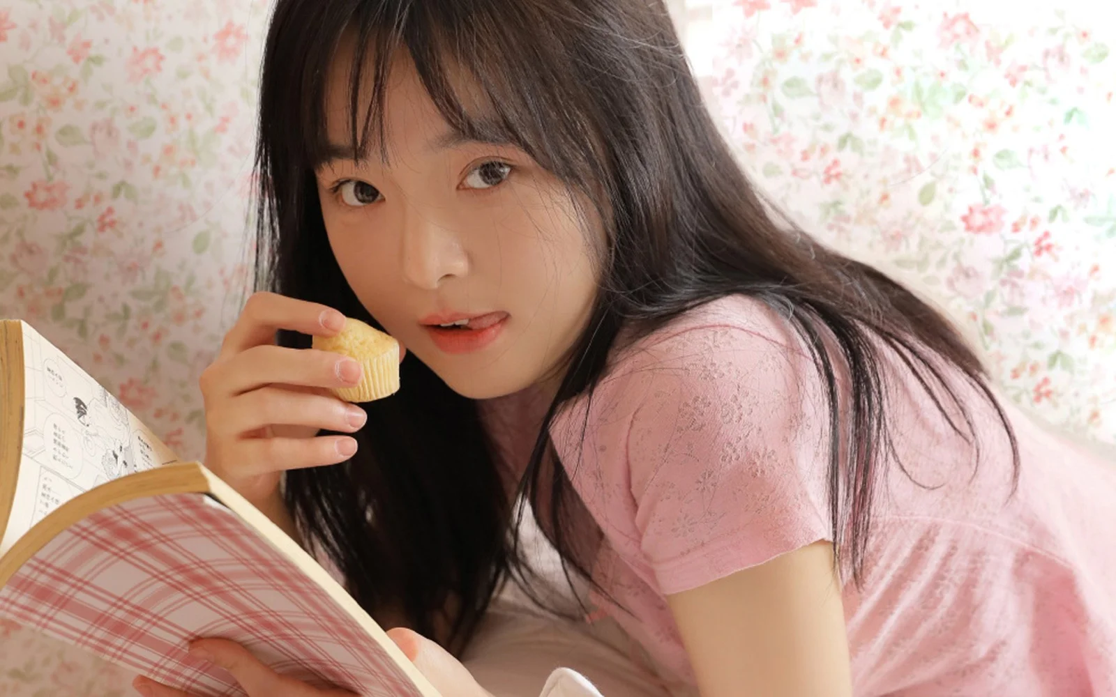一名年轻女子穿着粉色上衣，手拿小点心靠在书本旁，背景是花卉图案墙纸。