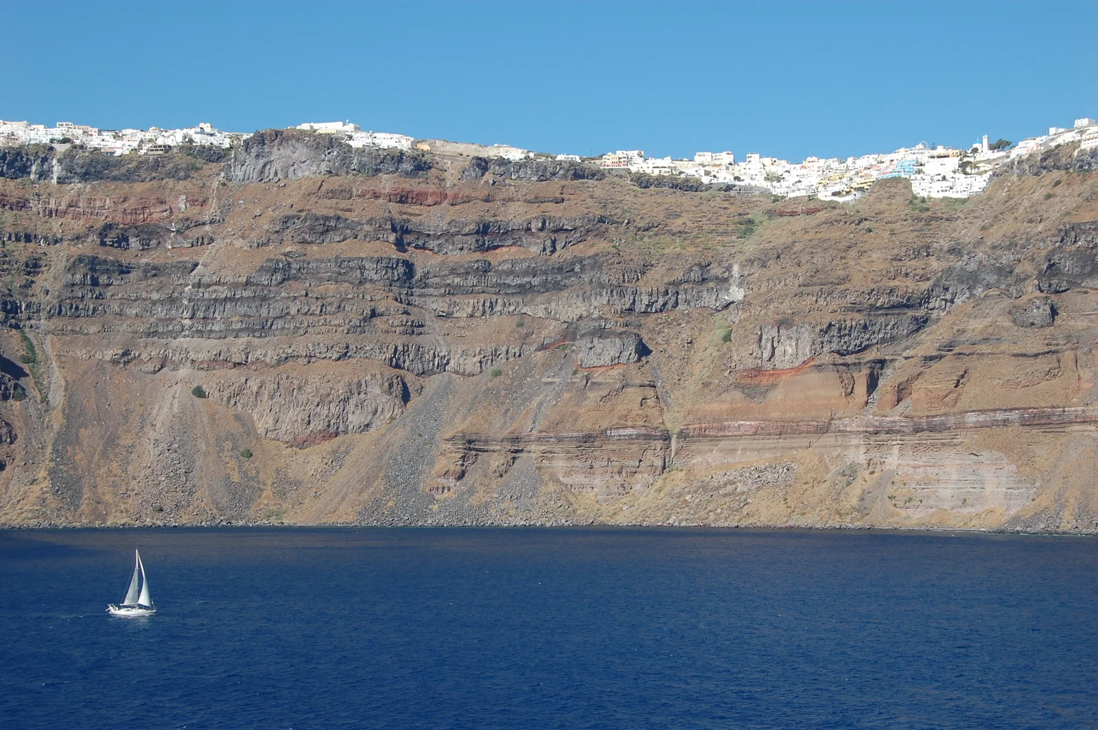蓝色海面前是层叠的火山悬崖，崖顶分布着白色建筑，海上有一艘小帆船。