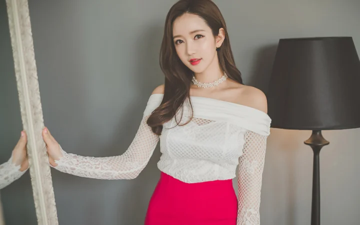 一名成年女性身穿白色上衣站在室内镜子和落地灯旁。