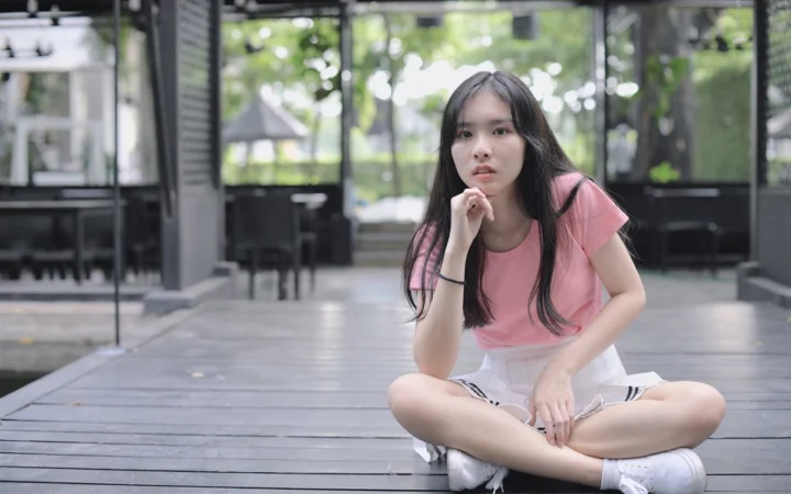 一名穿粉色上衣和白色短裙的女性坐在木质平台上，背景是明亮的户外休闲空间。