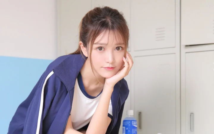 一名年轻女性穿着深蓝色外套，托腮坐在室内看向镜头。
