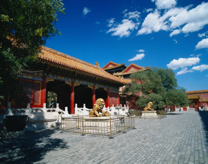 蓝天下的古典宫殿建筑环绕着铺满石砖的庭院，前方摆放着金色狮子雕像。