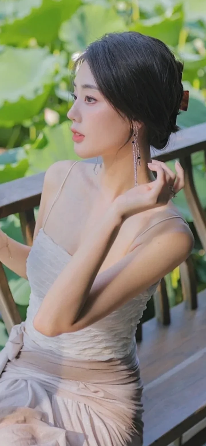 一位穿着浅色连衣裙的女子在荷叶旁侧身整理头发。