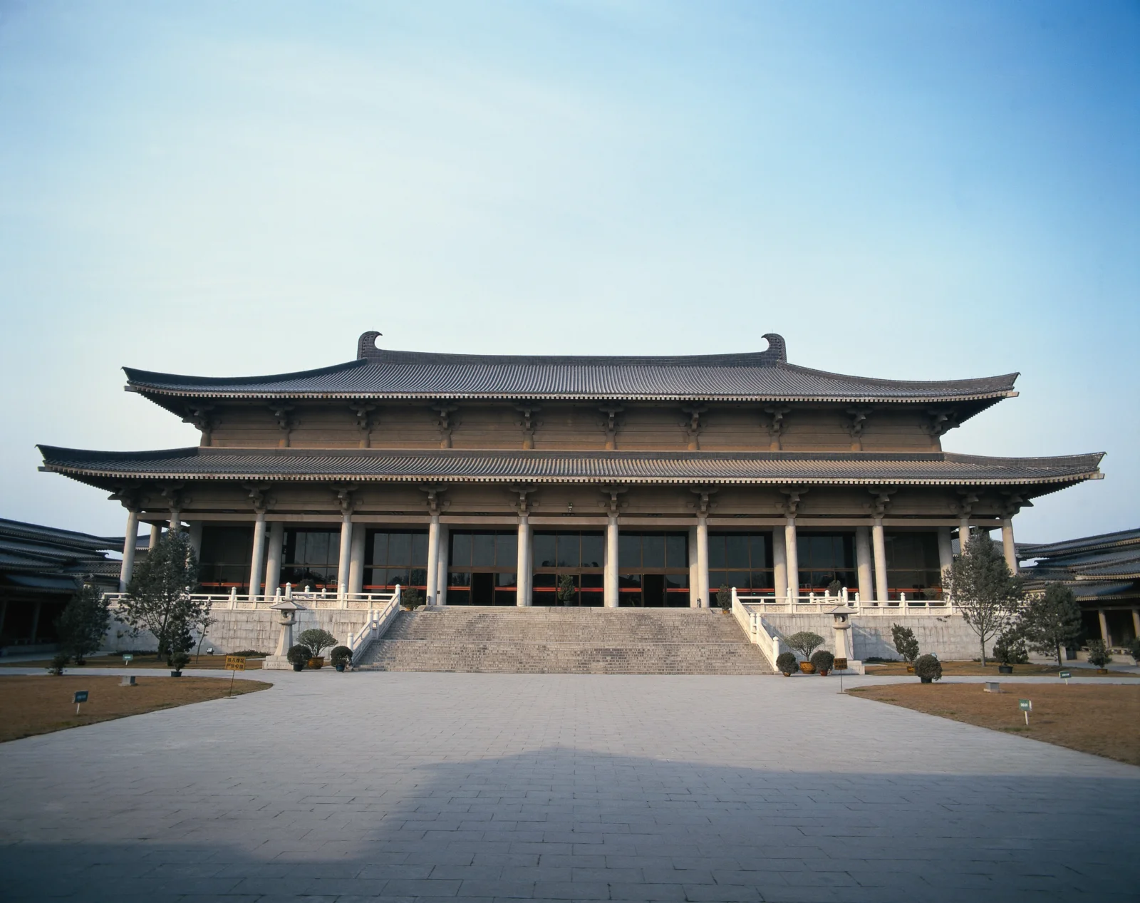 一座大型传统寺院建筑坐落在开阔的石板庭院前，背景是晴朗的浅蓝天空。