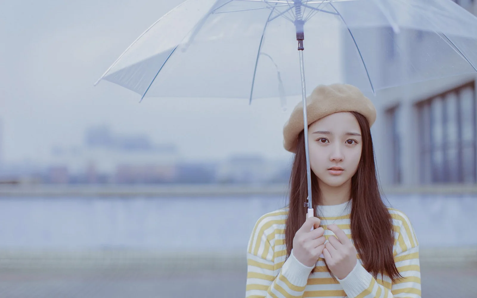 ベレー帽をかぶった女性が水辺で透明な傘を持っている。