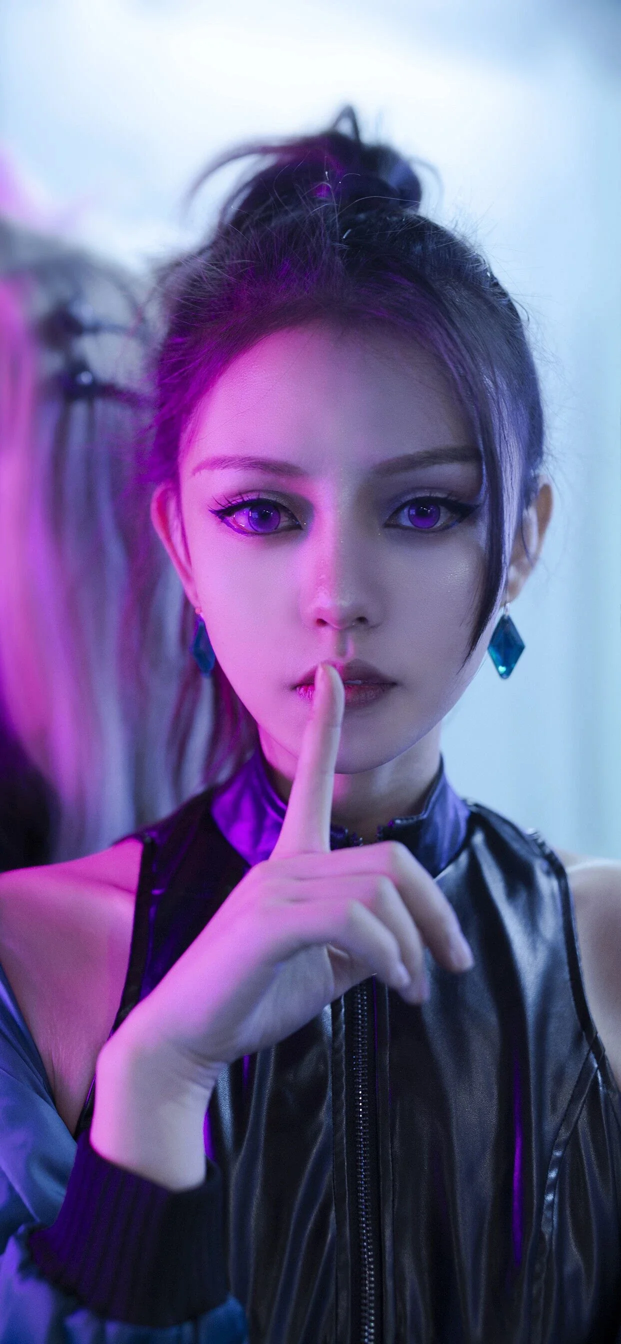 검은 가죽 재킷을 입은 성인 여성이 보라색과 파란색 조명 아래에서 카메라를 향해 쉿 하는 손짓을 하고 있다.