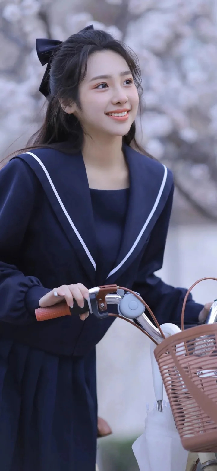 一名身穿深色水手风服装的年轻女子在花树下扶着一辆带篮子的自行车。