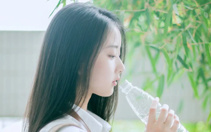 一位女性在窗边手持透明水瓶饮水，身旁可见绿色竹叶。