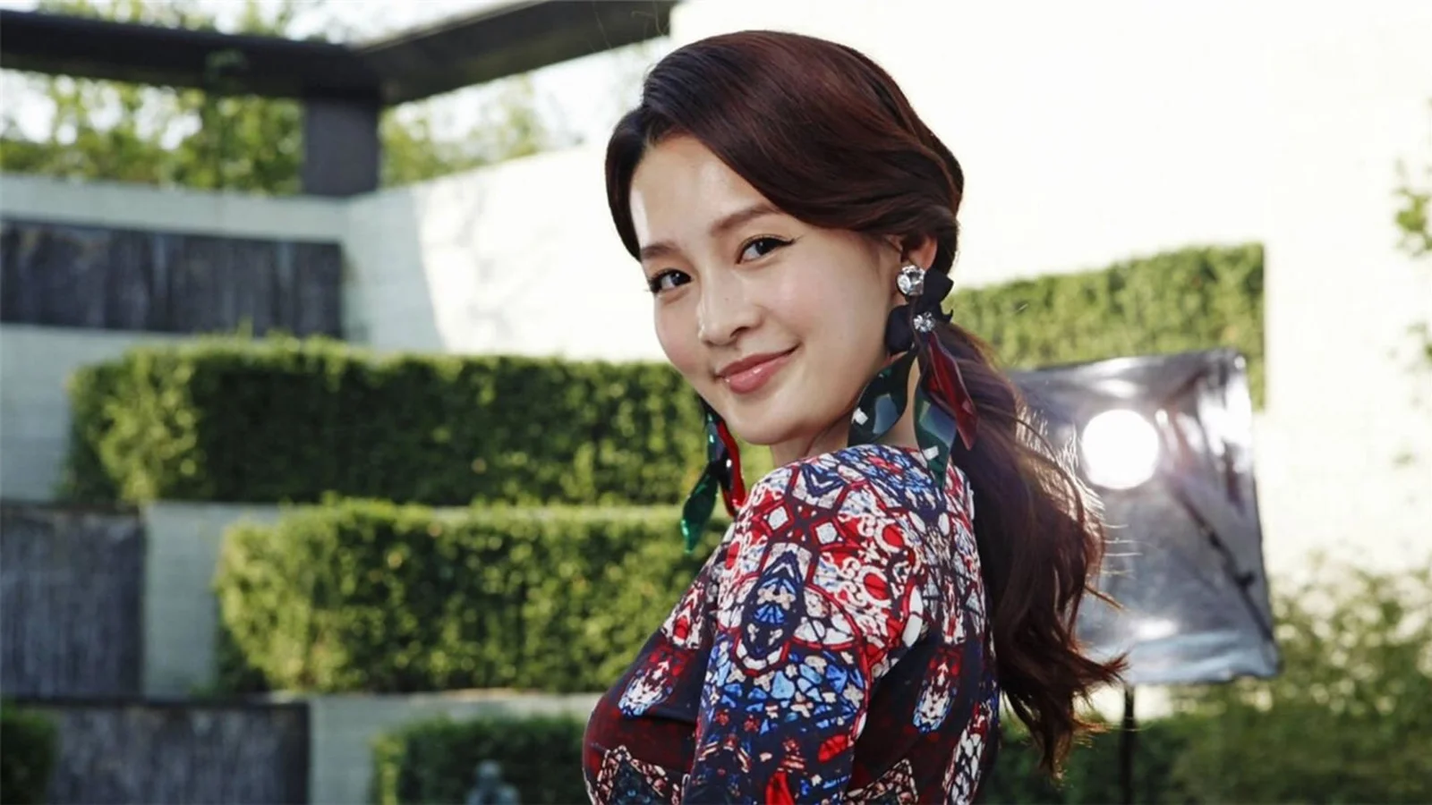 화려한 무늬의 옷을 입은 성인 여성이 깔끔하게 다듬어진 생울타리 앞에 서 있습니다.