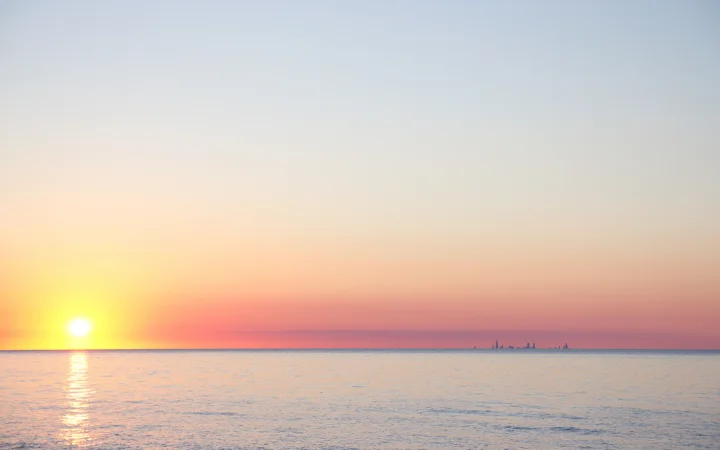 夕阳贴近海平线，金色光芒映照着平静的海面，远处隐约可见城市天际线。
