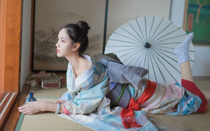 一名身穿传统服饰的女子趴在室内榻榻米上，身后放着一把白色纸伞。