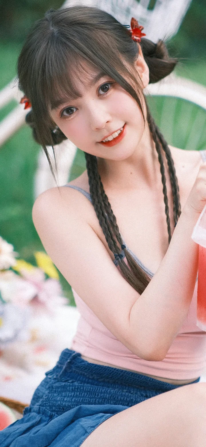 一位留着双辫、身穿粉色上衣的女性在户外花草背景前微笑。