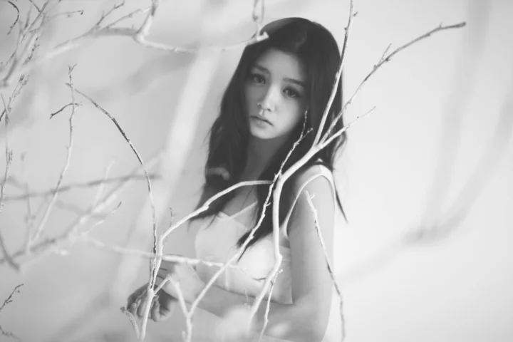 一位成年女性站在细枝之间，以黑白影调呈现柔和的人像画面。