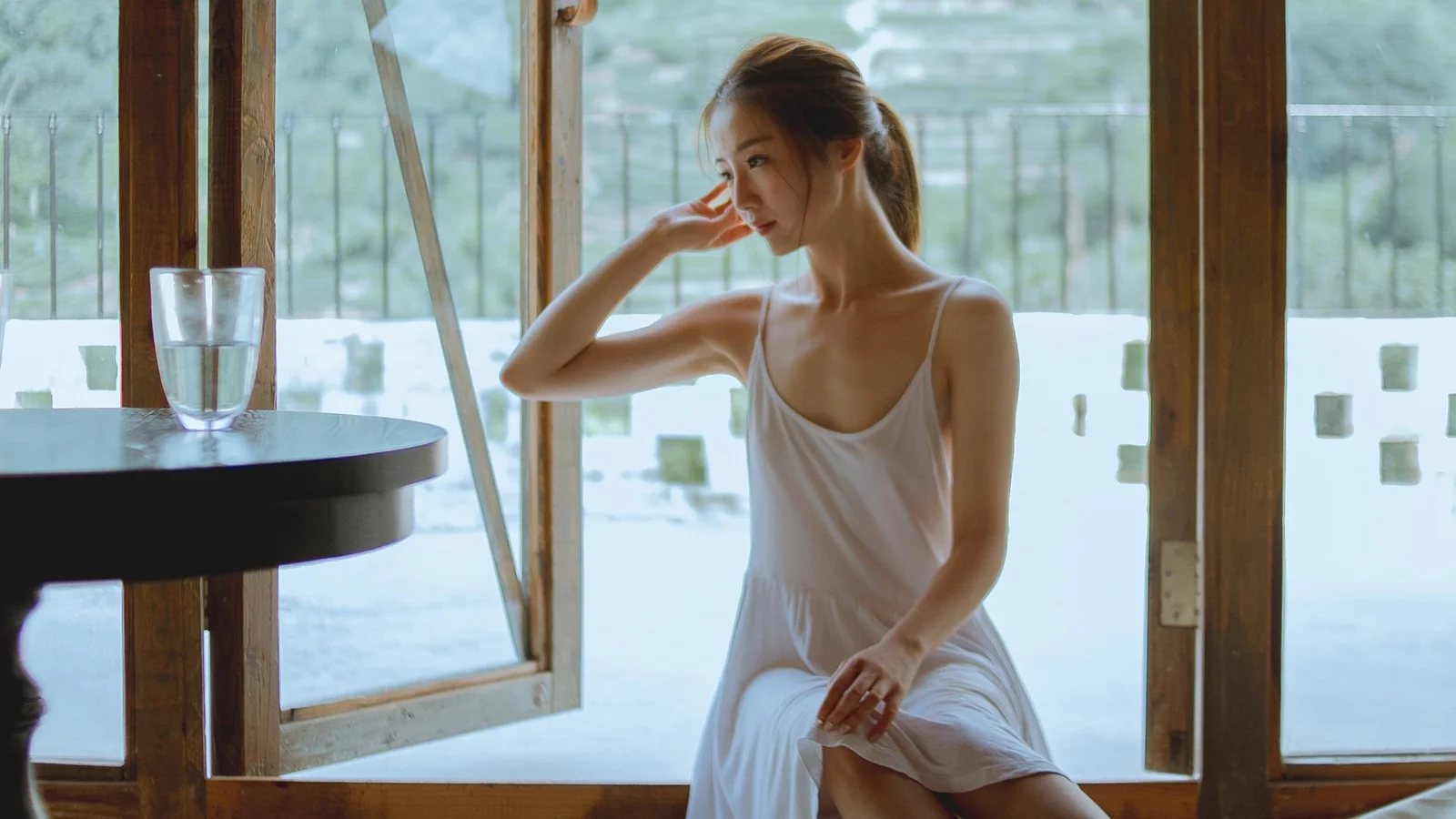 一名身穿白色連衣裙的女子坐在木質窗邊，窗外映入明亮的自然光。