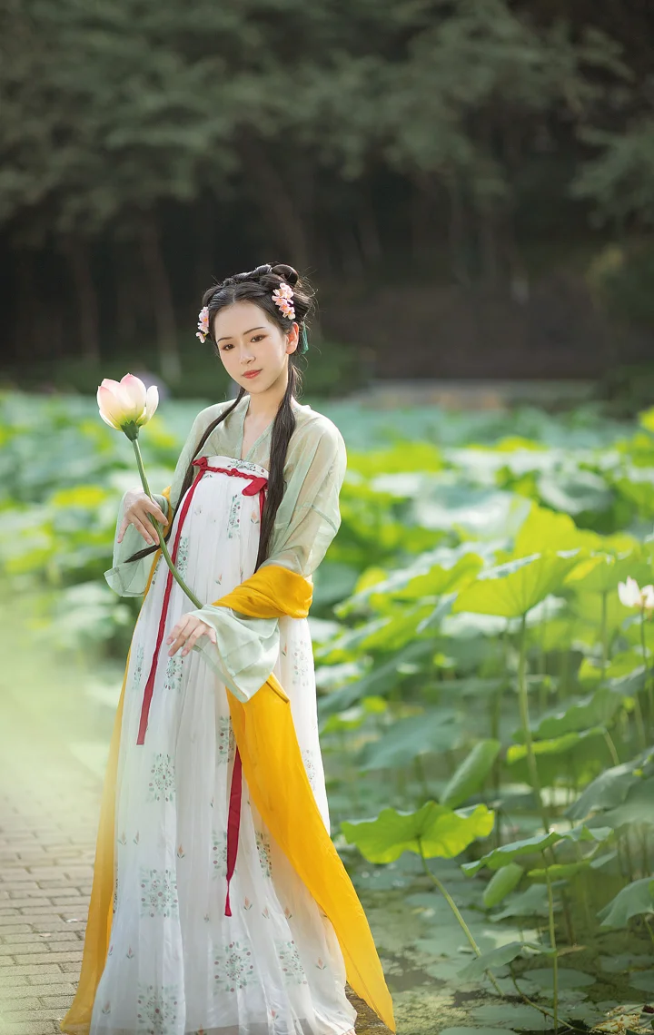 一位身穿浅色传统风格长裙的成年女子站在荷塘旁，手持一朵荷花。