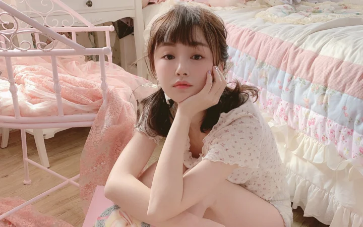 一名短发女性坐在粉色卧室的床边，手托着脸颊看向镜头。