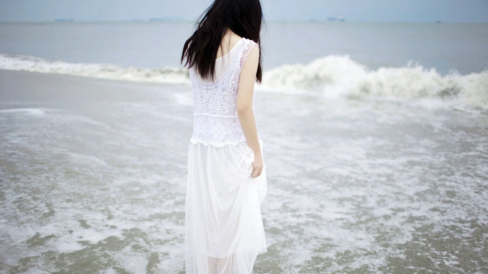 一名身穿白色连衣裙的女子站在海浪拍打的海滩上。
